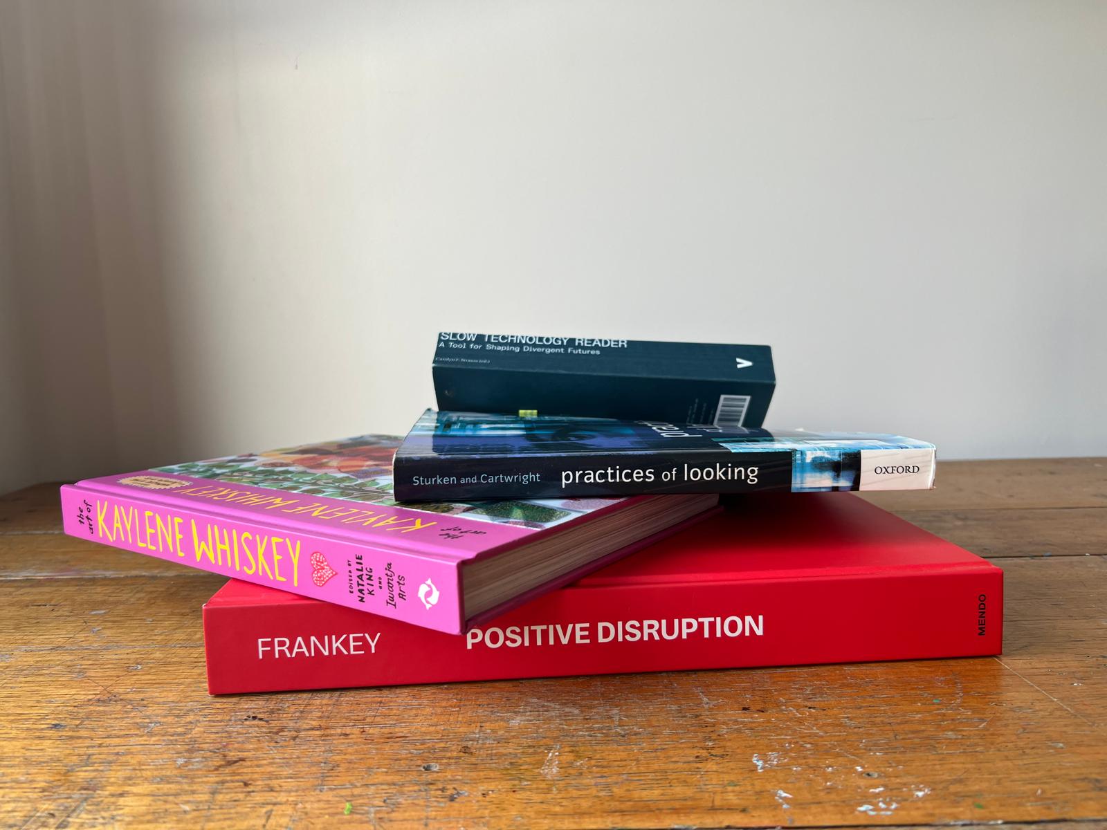 Four hardcover art and design books stacked at slightly different angles on a worn wooden studio table, their edges running in different directions.