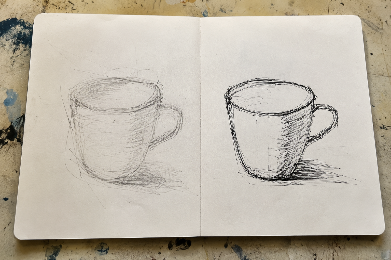Two observational drawings of the same cup in an open sketchbook. The graphite drawing contains light searching and corrected lines, while the fineliner drawing contains darker repeated marks that remain visible.