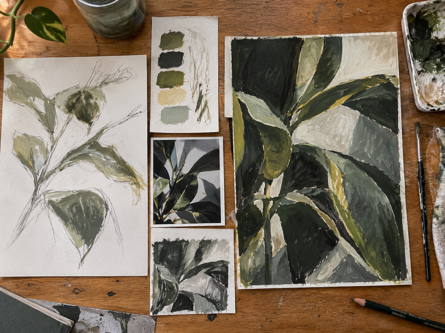 Studio process laid out on a wooden table: an earlier plant study, a more developed version, a source photograph, smaller crop and compositional studies, and colour and material tests, showing visible changes between stages.
