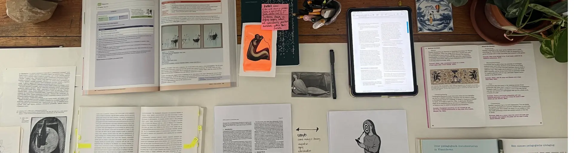 Overhead view of an artistic research workspace: open books, printed articles, drawings, handwritten notes, sticky notes and a tablet arranged across a desk