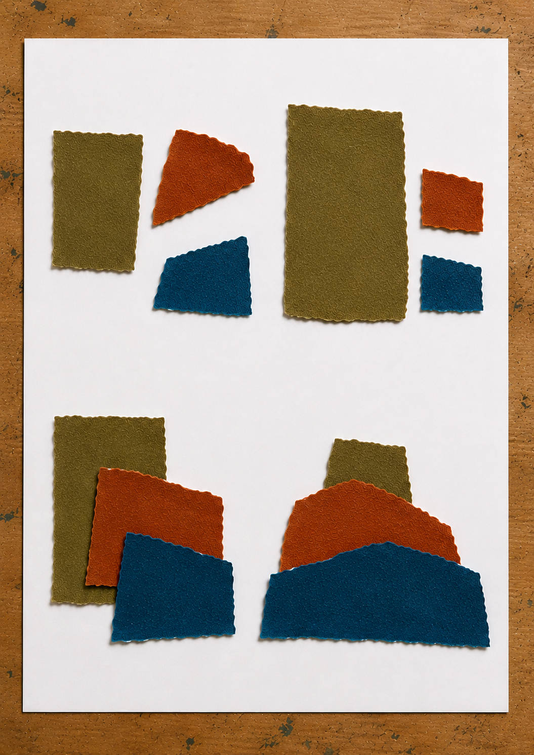 Torn paper studies on a white sheet of paper resting on a wooden desk: four arrangements exploring position, size, overlap and depth using olive, rust and blue textured paper fragments.
