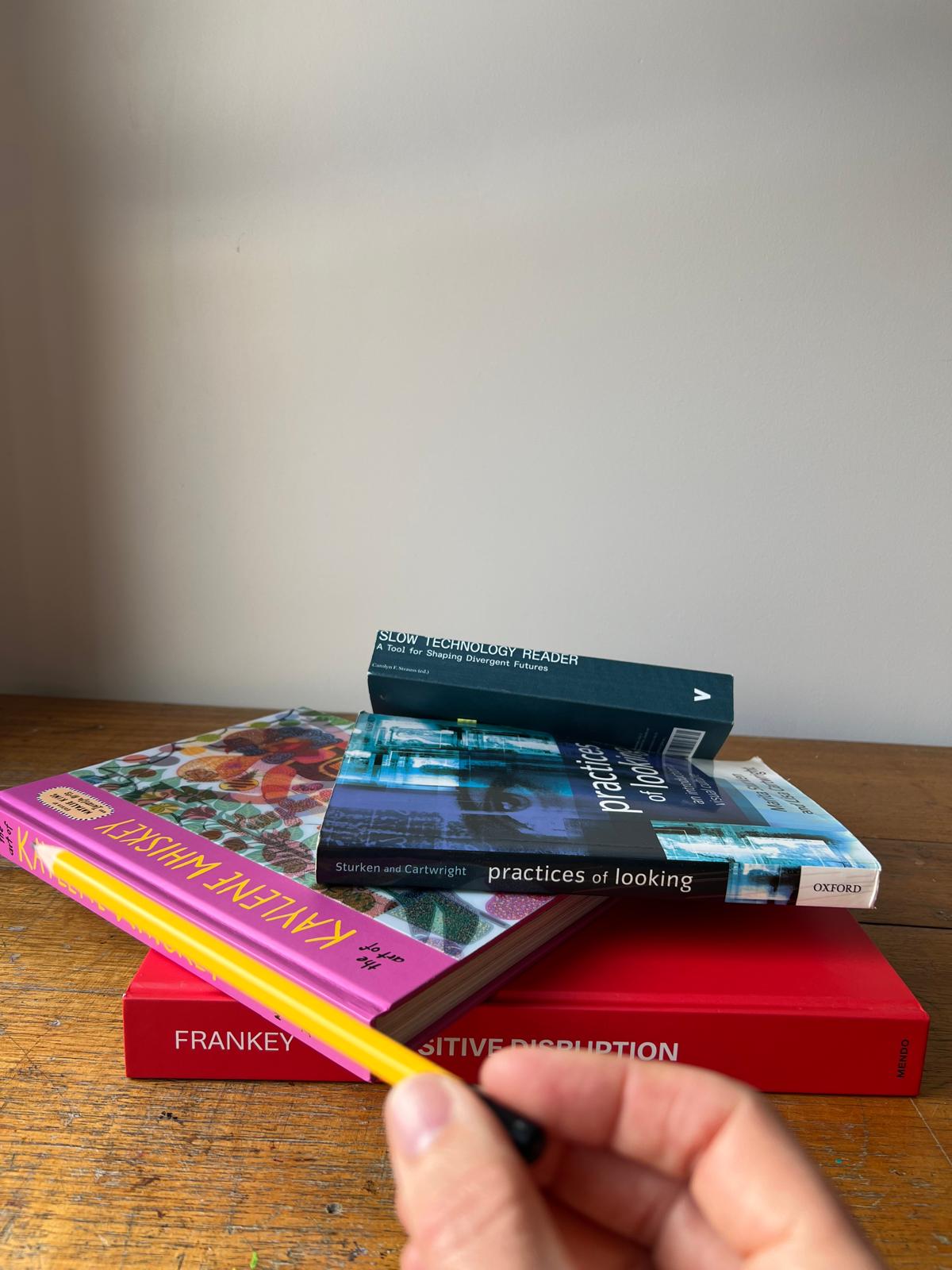 A hand holding a yellow pencil at arm's length, the pencil lined up with the receding top edge of a pink hardcover book in a stack of books on a studio table.