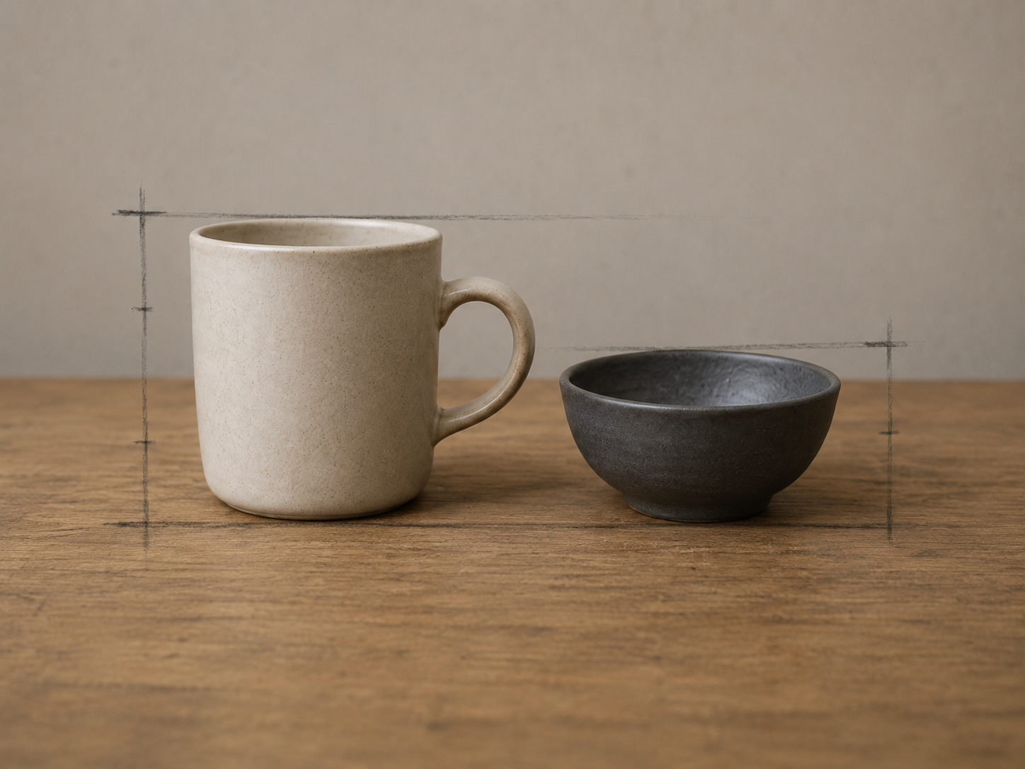 A cream stoneware mug and a smaller dark stoneware bowl standing side by side on a worn wooden table against a plain grey wall, with faint hand-drawn graphite comparison marks in the air around them.