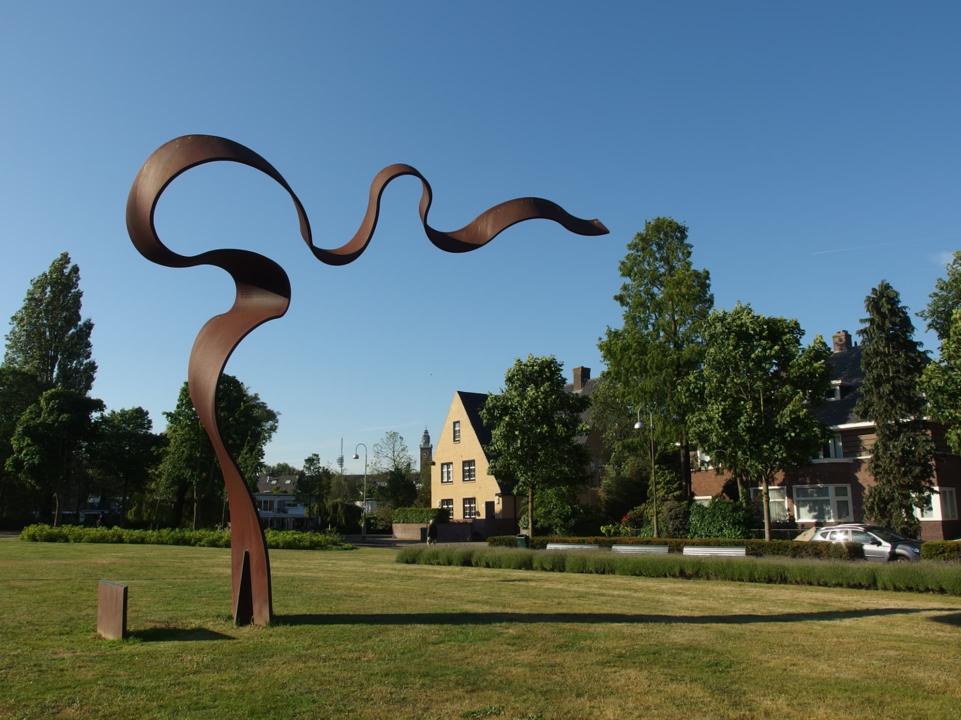 A tall curving steel sculpture standing on grass beside a street in Amsterdam Zuid, with trees, houses and parked cars behind it.